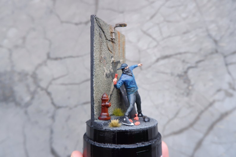 May include: A miniature figurine of a person wearing a blue jacket and black pants spray painting a wall. The wall is made of gray concrete and has a red fire hydrant at the base. The figurine is standing on a black base.