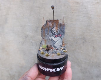 May include: A miniature diorama of a graffiti art piece by Banksy. The diorama features a white cat with a red bow tie painted on a crumbling brick wall. The diorama is set on a black base with the word "Banksy" printed on it.