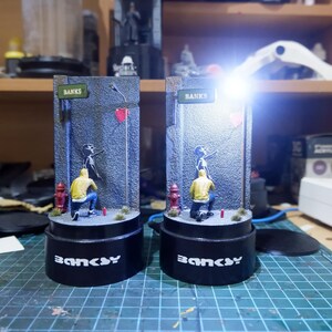 May include: Two black and white Banksy-style street art figurines with a red heart and the word "Banksy" on the base. The figurines are on a black base with a white background.
