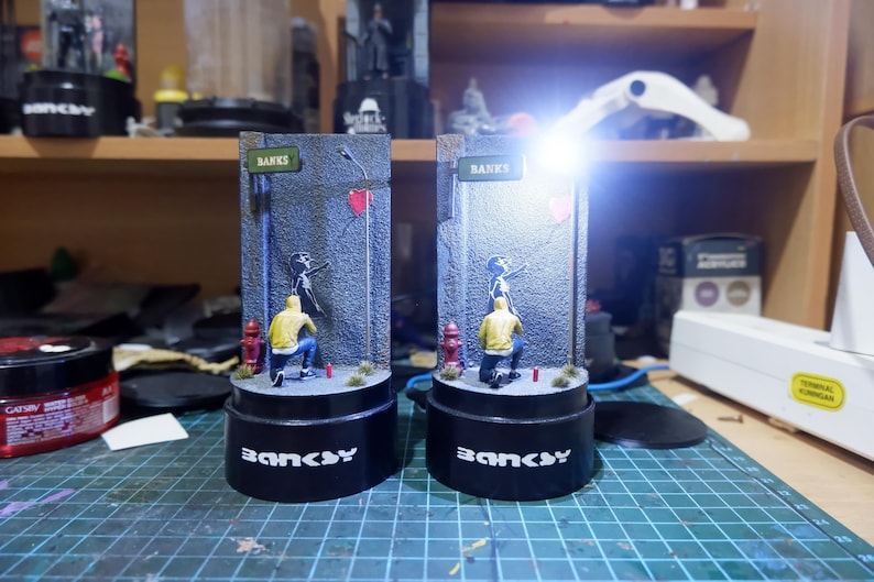 May include: Two black and white Banksy-style street art figurines with a red heart and the word "Banksy" on the base. The figurines are on a black base with a white background.