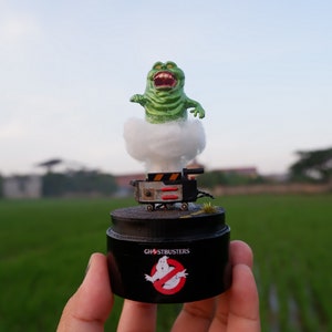 Puede incluir: Una figura verde de los Cazafantasmas con una base de nube blanca, sentada sobre una base negra con el logotipo de los Cazafantasmas. La figura sostiene un pequeño dispositivo negro y rojo.