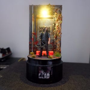 Puede incluir: Diorama en miniatura con una figura de "The Walking Dead" frente a una pared desgastada con luz. La base negra muestra el t&iacute;tulo de la serie. La escena incluye una ballesta y follaje.