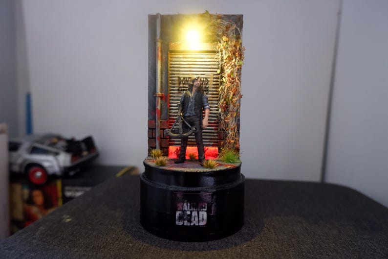 Puede incluir: Diorama en miniatura con una figura de "The Walking Dead" frente a una pared desgastada con luz. La base negra muestra el t&iacute;tulo de la serie. La escena incluye una ballesta y follaje.