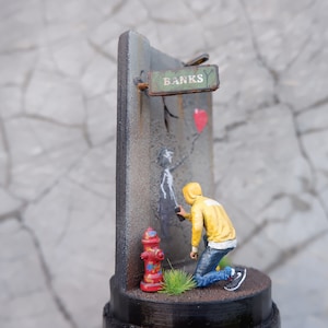 May include: A miniature diorama depicting a street scene with a figure painting a stencil on a wall. The stencil is of a person holding a red heart balloon. The wall has a sign that reads "Banks" and a fire hydrant is in the foreground.