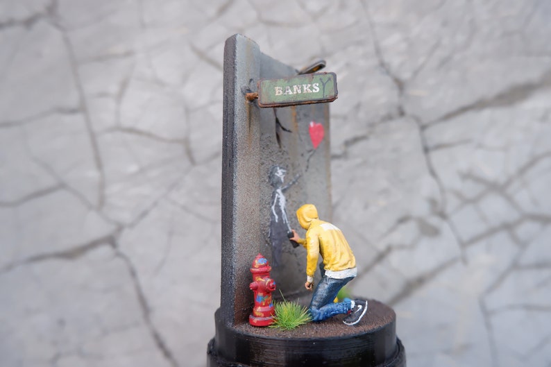 May include: A miniature diorama depicting a street scene with a figure painting a stencil on a wall. The stencil is of a person holding a red heart balloon. The wall has a sign that reads "Banks" and a fire hydrant is in the foreground.