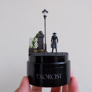 May include: A miniature diorama of a man in a black coat and hat standing in front of a street lamp and a stone wall with a chain link fence. The diorama is on a black base with the word "EXORCIST" printed on it.