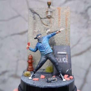 May include: A miniature figure of a person wearing a blue jacket, black pants, and a black bandana over their face. The figure is holding a red object in their right hand and is throwing it towards a wall with a cracked surface. The wall has a sign that says "POLICE" on it. The figure is standing on a small, black base.