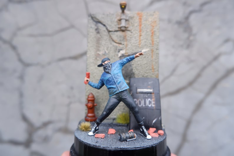 May include: A miniature figure of a person wearing a blue jacket, black pants, and a black bandana over their face. The figure is holding a red object in their right hand and is throwing it towards a wall with a cracked surface. The wall has a sign that says "POLICE" on it. The figure is standing on a small, black base.