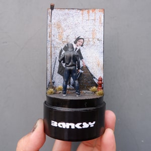 May include: A miniature diorama of a street scene featuring a hooded figure spray painting a mural of a woman in a maid's uniform. The diorama is on a black base with the word "Banksy" printed on it.