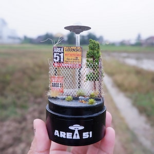 Puede incluir: Diorama en miniatura del Área 51 con una base negra con "AREA 51" y un gráfico de OVNI. La escena incluye una valla de alambre, señales de advertencia y un pequeño árbol, coronado por un OVNI brillante.