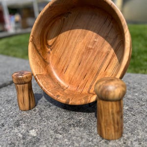 May include: A wooden bowl and two matching salt and pepper shakers. The bowl is large and round, with a natural wood grain pattern. The shakers are cylindrical with rounded tops, also made of wood.