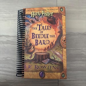 May include: A spiral-bound book titled The Tales of Beedle the Bard by J.K. Rowling. The cover features a colorful illustration of a cauldron, a tree stump with an axe, and magical imagery in shades of orange, brown, and purple with gold accents.