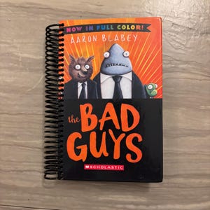 May include: A spiral-bound book titled "The Bad Guys" by Aaron Blabey. The cover features illustrations of anthropomorphic characters, including a wolf and a shark, in suits. The title is in large orange letters against a black background.
