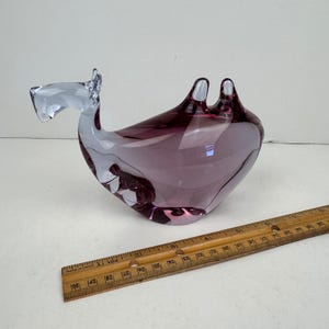 May include: A clear and deep purple glass camel figurine with two humps, positioned next to a wooden ruler that shows the object is approximately 10 inches in length. The glass sculpture features a smooth, curved body and a distinct head shape.