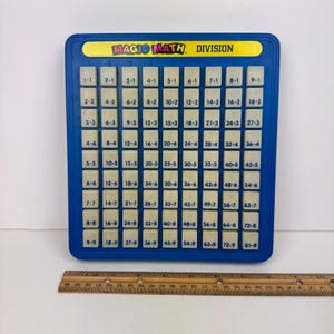 May include: A blue Magic Math Division board game with a yellow label. The board has a grid of white tiles with multiplication problems. A wooden ruler is at the bottom. The game is designed for educational purposes.
