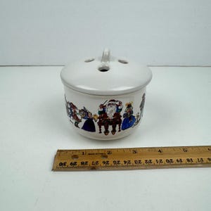 Op de afbeelding: Witte keramische kersttheedoos met deksel. De doos heeft een band met kleurrijke illustraties van Sinterklaas en elven die muziek maken. Het deksel heeft een klein handvat.