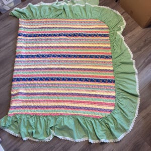 May include: A rectangular quilt featuring horizontal stripes of floral, gingham, and solid yellow patterns. The quilt is bordered by a wide, green and white gingham ruffle trimmed with white lace along the entire outer edge of the fabric.