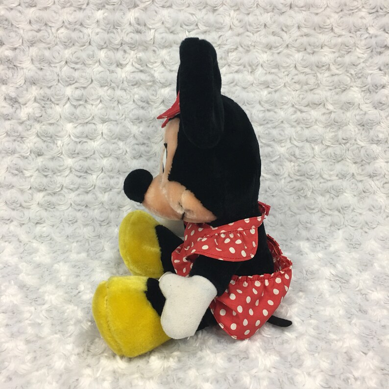 May include: A plush toy of Minnie Mouse wearing a red and white polka dot dress with a yellow bow. She is sitting with her legs crossed and her arms resting on her lap.