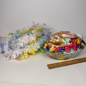 May include: An Easter-themed ceramic basket with a bunny and floral design. The basket is decorated with colorful flowers, a red bow, and small yellow chicks. A garland with pastel-colored butterfly cutouts is next to the basket.