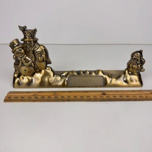 Könnte beinhalten: Eine bronzefarbene Weihnachtsdekoration mit zwei Schneemännern links und dem Weihnachtsmann rechts. Die Schneemänner tragen Zylinder und Schals. Die Basis hat einen rechteckigen Bereich zur Personalisierung.