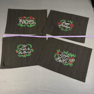May include: Four dark brown placemats with embroidered holiday greetings. Each mat features a different phrase: "NOEL", "Happy Holidays", "Eat Drink & Be Merry", and "Jingle Bells", surrounded by festive wreaths.