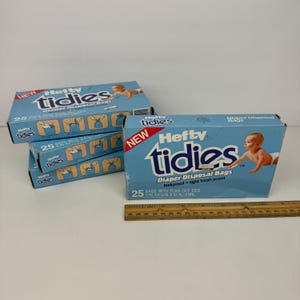 Puede incluir: Varias cajas de bolsas para desechar pañales Hefty Tidies están apiladas sobre una superficie blanca. Las cajas son de color azul claro con el nombre de la marca en blanco y un bebé. Cada caja contiene 25 bolsas.