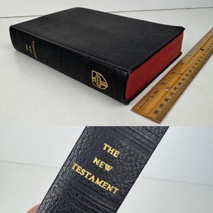 May include: A black leather-bound book with red page edges. The spine reads "THE NEW TESTAMENT" in gold lettering. A wooden ruler is next to the book. The book has a gold emblem on the front cover.