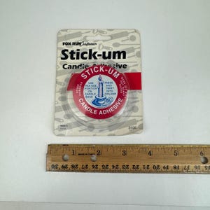 Può includere: Un pacchetto di adesivo per candele Stick-um, un adesivo trasparente per fissare le candele ai portacandele. La confezione è rossa e bianca con un'immagine di una candela e un portacandele. Il testo sulla confezione recita "Stick-um Candle Adhesive" e "Never Hardens".