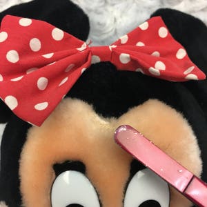 May include: A plush toy of a cartoon mouse with black ears and a red bow with white polka dots. The mouse has a pink nose and large black eyes.