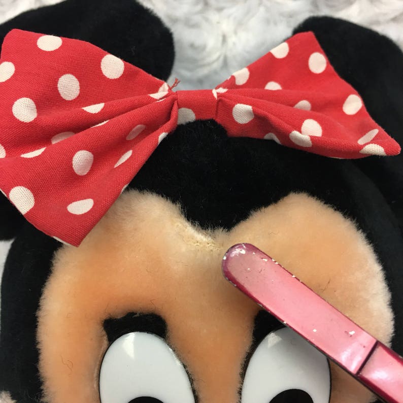 May include: A plush toy of a cartoon mouse with black ears and a red bow with white polka dots. The mouse has a pink nose and large black eyes.