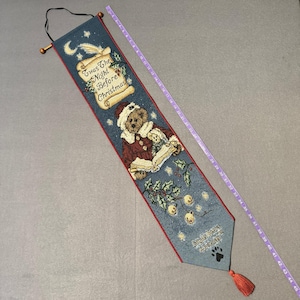 May include: A Christmas tapestry with a blue background, red trim, and a tassel. It features a teddy bear in a Santa hat reading a book with the text "Twas The Night Before Christmas". The tapestry is approximately 45 inches long.