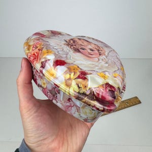 May include: Heart-shaped tin box with a floral pattern and an angel illustration on the lid. The box features a mix of pink, yellow, and white flowers. The box is held in a hand.
