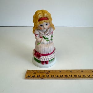 Puede incluir: Una figurita de porcelana de una niña con cabello rubio que lleva un vestido rosa y zapatos rojos. Ella sostiene un ramo de flores blancas y está sentada sobre una base verde.