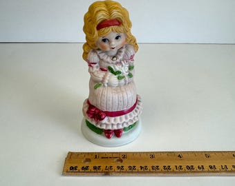 May include: A porcelain figurine of a girl with blonde hair wearing a pink dress and red shoes. She is holding a bouquet of white flowers and is sitting on a green base.