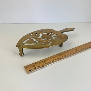 May include: A brass trivet in the shape of a heart with a floral design. The trivet has three legs and a handle.