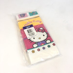 May include: A package of Hello Kitty party table covers. The covers are white with a pink, yellow, and green border. The center of the cover features a large image of Hello Kitty with the words "Hello Kitty" printed above her. The package also includes a small sticker with the words "Paper Art" and a barcode.