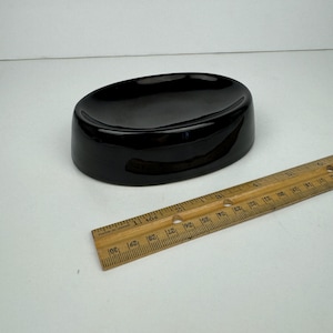 May include: A glossy black ceramic soap dish with an oval shape. The dish is approximately 5 inches long and 3 inches wide. A wooden ruler is next to the dish.