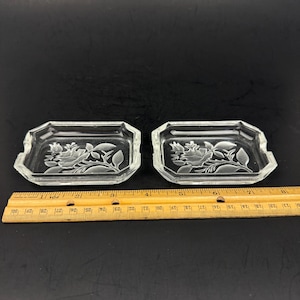 Puede incluir: Dos platos de vidrio transparente con un diseño floral. Cada plato es rectangular con forma octogonal y un patrón floral en relieve. Una regla de madera se coloca debajo.