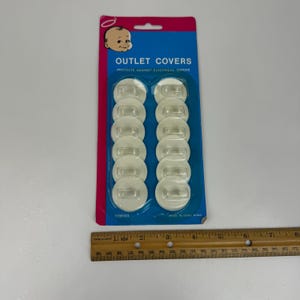 Puede incluir: Un paquete de protectores de enchufes de plástico transparente. El paquete es azul y rosa con las palabras "OUTLET COVERS" y un gráfico de bebé. Los protectores están diseñados para proteger contra descargas eléctricas.