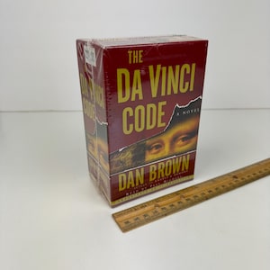 May include: A boxed copy of Dan Brown's novel, "The Da Vinci Code," in a maroon box with gold lettering. The book cover features an image of eyes and a torn paper effect. A wooden ruler is next to the book.