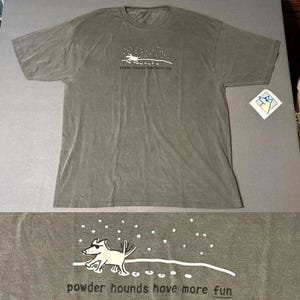 May include: A dark gray t-shirt with a graphic of a dog wearing sunglasses, running on a snowy hill. The text "powder hounds have more fun" is printed below the graphic. The shirt is made of a soft material.