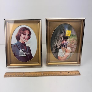 May include: Two gold-colored oval picture frames, each holding a 5x7 inch photo. The frames have a metallic finish and a clear glass cover. A wooden ruler is placed below the frames.