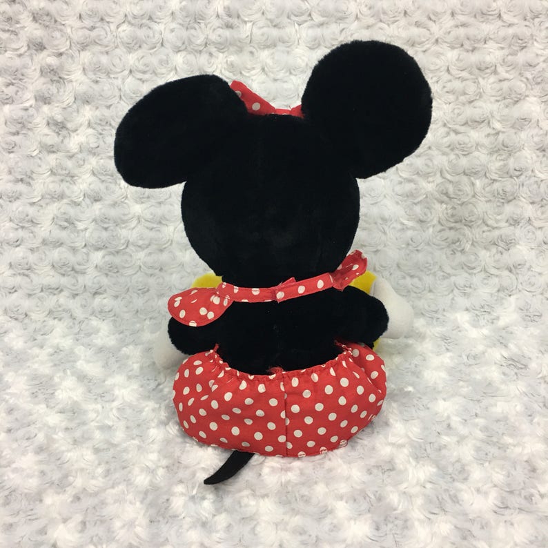 May include: Black and white plush toy of a cartoon mouse wearing a red and white polka dot dress and a red bow. The toy is sitting on a white fluffy surface.