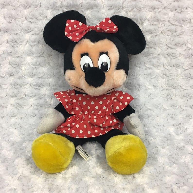 May include: A plush Minnie Mouse toy wearing a red dress with white polka dots and a red bow with white polka dots. The toy has yellow shoes and a black nose.