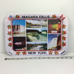 May include: A white metal tray with a red, white, and blue American flag and a Canadian flag on either side. The tray features a collage of images of Niagara Falls, including Horseshoe Falls, Prospect Point, and Niagara Falls illuminated at night. The tray also includes the text "NIAGARA FALLS" and "NEW YORK".