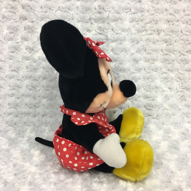 May include: A plush Minnie Mouse toy wearing a red and white polka dot dress with a red bow. She has black ears and a black nose. Her feet are yellow.