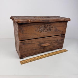 Peut inclure: Une boîte à bijoux en bois marron avec deux tiroirs et un motif de tournesol sculpté sur le devant. La boîte a un aspect texturé de grain de bois. Une règle en bois est placée à côté de la boîte. La règle mesure environ 30 cm.