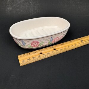 May include: An oval ceramic soap dish with a cream-colored interior featuring raised ridges. The exterior has a white lattice pattern decorated with painted pink roses and blue flowers. A wooden ruler shows the dish is approximately 6 inches long.