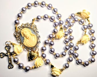 Puede incluir: Un collar de rosario plateado y dorado con un colgante de camafeo que presenta un retrato de Jesús. Las cuentas del rosario están intercaladas con acentos de rosas blancas.