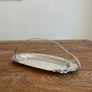 May include: An oval silver-toned metal serving tray featuring an arched, slender handle. The tray has a smooth, reflective central surface and decorative, ornate scrollwork detailing along the edges and the base of the handle attachment points.